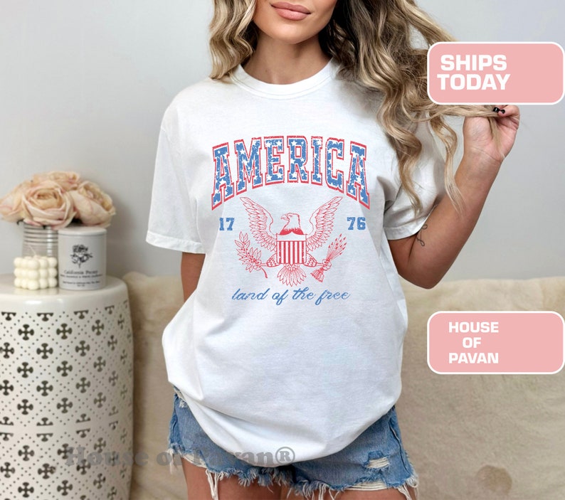 May include: White t-shirt with a vintage-style graphic of an eagle with the words "AMERICA 17 76 land of the free" printed on it.
