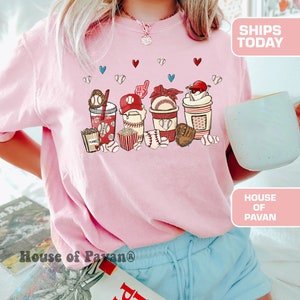 May include: Pink t-shirt with a graphic of baseball-themed drinks, popcorn, and baseballs. The graphic includes the words "PLANNER" and "HOME".