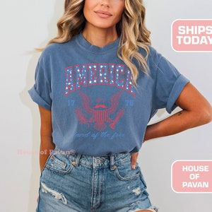 May include: A blue t-shirt with a vintage-style graphic of an eagle and the text "America 1776 Land of the Free".