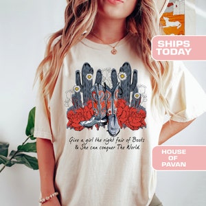 Op de afbeelding: Een beige T-shirt met een afbeelding van cowboylaarzen, cactussen en bloemen. De tekst "Give a girl the right pair of Boots & She can conquer The World" staat op het T-shirt gedrukt.