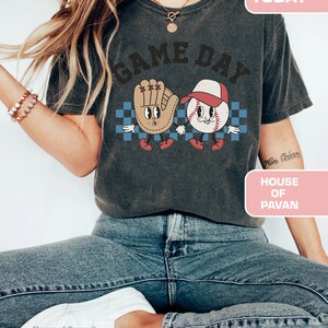 以下が含まれることがあります： 「GAME DAY」という文字がプリントされたグレーのTシャツ。Tシャツには、漫画の野球ボールとグローブが手をつないでいる様子が描かれている。「HOUSE OF PAVAN」という文字が、画像の右下隅にあるピンク色の長方形にプリントされている。