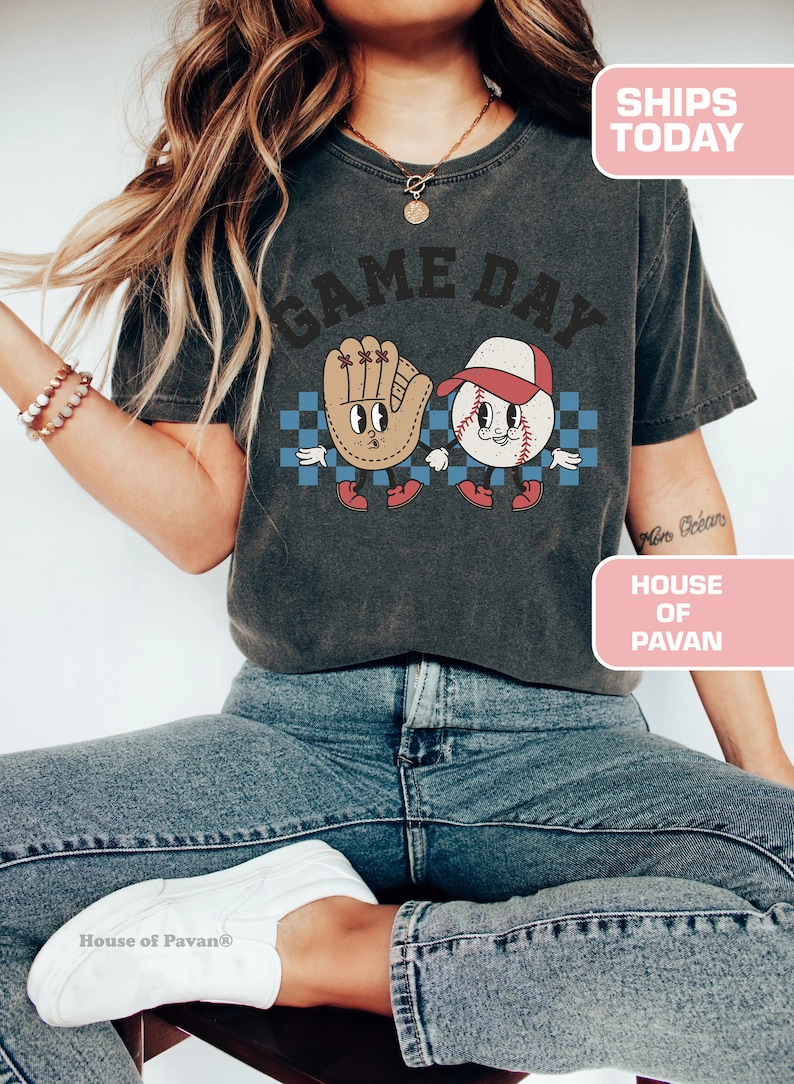 以下が含まれることがあります： 「GAME DAY」という文字がプリントされたグレーのTシャツ。Tシャツには、漫画の野球ボールとグローブが手をつないでいる様子が描かれている。「HOUSE OF PAVAN」という文字が、画像の右下隅にあるピンク色の長方形にプリントされている。