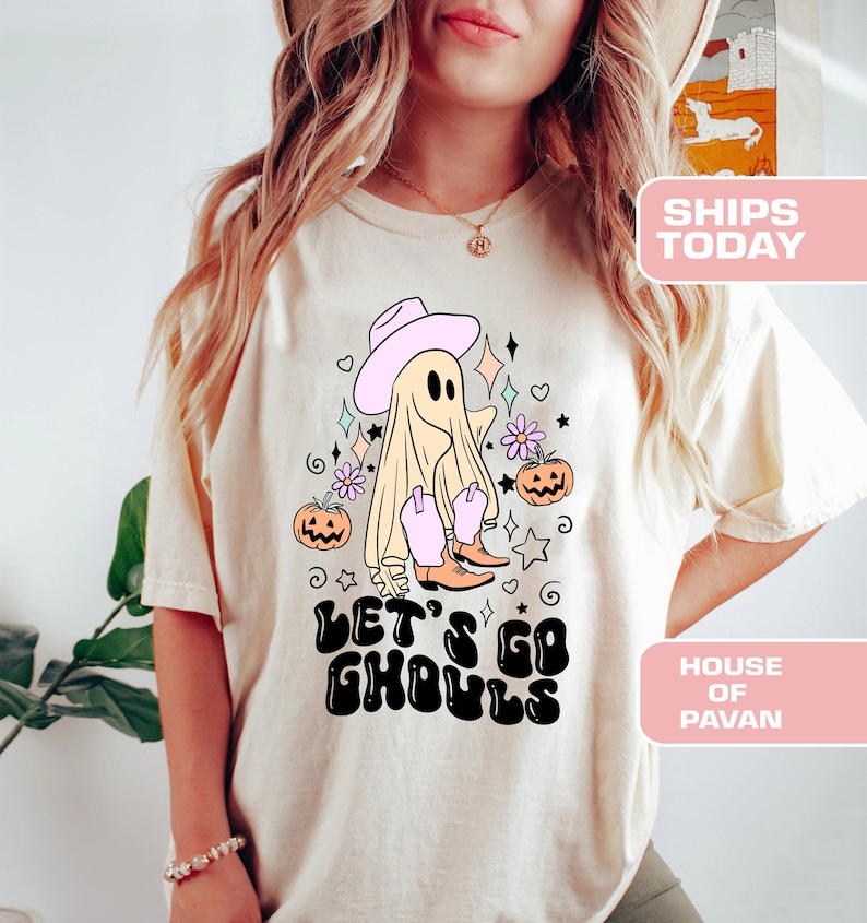 K&ouml;nnte beinhalten: Ein wei&szlig;es T-Shirt mit einem Cartoon-Geist, der einen rosa Cowboyhut und Stiefel tr&auml;gt. Der Geist ist von K&uuml;rbissen und Sternen umgeben. Der Text "Let's Go Ghouls" ist in Schwarz und Wei&szlig; auf das Shirt gedruckt.