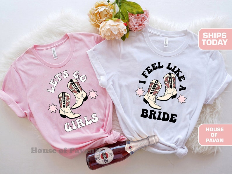 K&ouml;nnte beinhalten: Zwei rosa und wei&szlig;e T-Shirts mit Western-Motiv. Das rosa Shirt sagt "Let's Go Girls" mit einem Paar Cowboystiefeln. Das wei&szlig;e Shirt sagt "I Feel Like A Bride" mit einem Paar Cowboystiefeln.