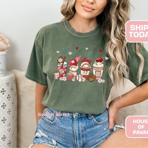 May include: A green t-shirt with a graphic of baseball-themed cups and snacks. The cups have red, white, and blue accents. The graphic includes baseballs, popcorn, and a baseball cap.