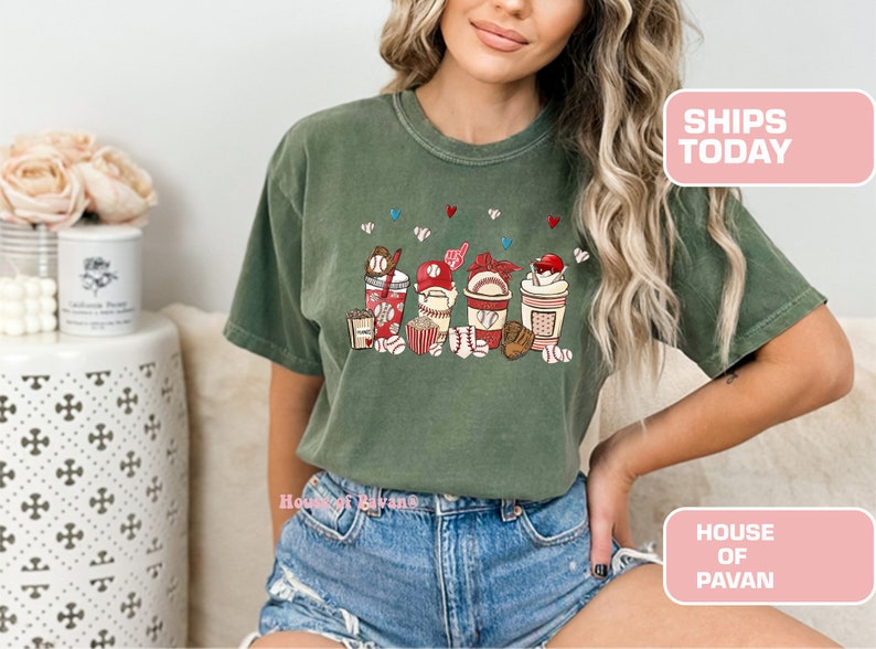 May include: A green t-shirt with a graphic of baseball-themed cups and snacks. The cups have red, white, and blue accents. The graphic includes baseballs, popcorn, and a baseball cap.