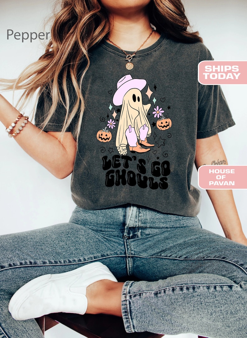 K&ouml;nnte beinhalten: Ein dunkelgraues T-Shirt mit einem Aufdruck eines Geistes, der einen rosafarbenen Cowboyhut und Stiefel tr&auml;gt. Der Geist ist von Sternen, Herzen und Blumen umgeben. Der Text "Let's Go Ghouls" ist in Schwarz unter dem Geist aufgedruckt.