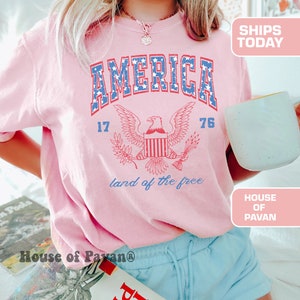 May include: A pink t-shirt with a vintage-style graphic of an eagle and the words "AMERICA 17 76 land of the free".