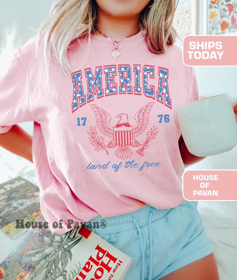 May include: A pink t-shirt with a vintage-style graphic of an eagle and the words "AMERICA 17 76 land of the free".