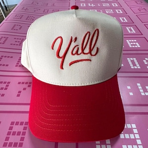 May include: A baseball cap featuring a cream-coloured crown and a red brim. The word "Y'all" is embroidered in red script across the front. The cap has a red button on top.