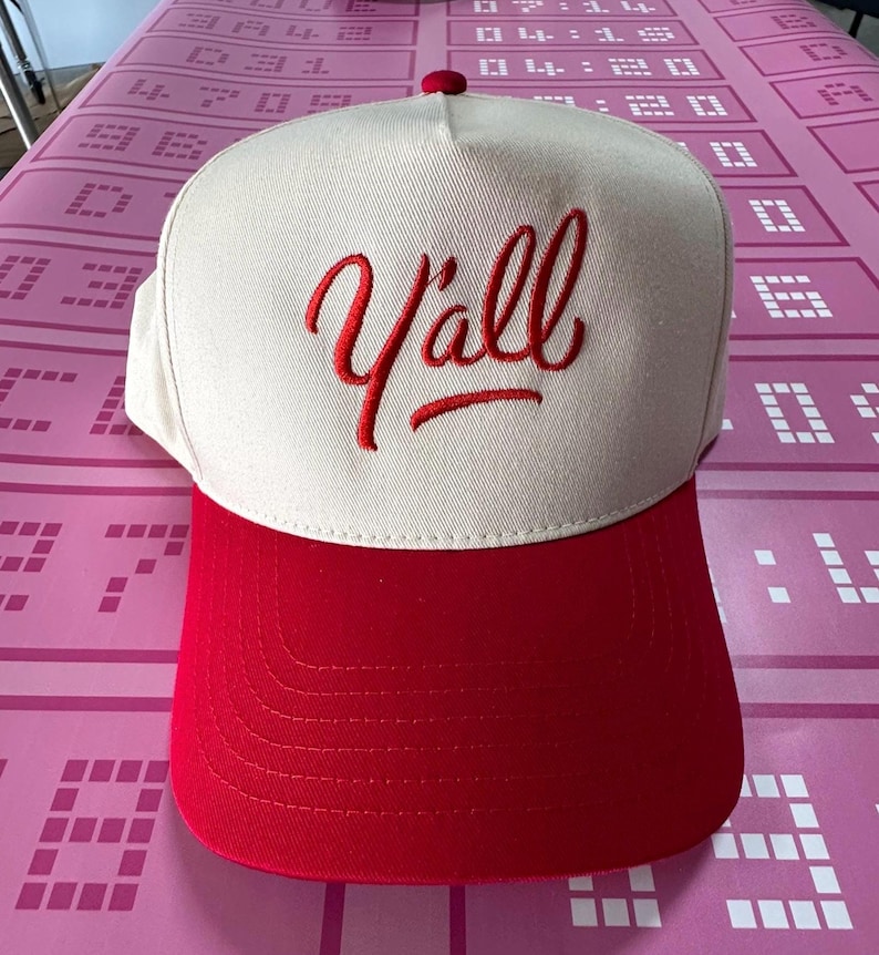 May include: A baseball cap featuring a cream-coloured crown and a red brim. The word "Y'all" is embroidered in red script across the front. The cap has a red button on top.