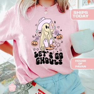 K&ouml;nnte beinhalten: Pinkes T-Shirt mit einem Cartoon-Geist, der einen Cowboyhut und Stiefel tr&auml;gt. Der Geist ist von Blumen und K&uuml;rbissen umgeben. Der Text "Let's Go Ghouls" ist in einer Retro-Schrift geschrieben.