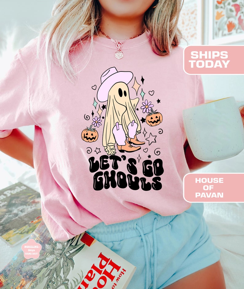 K&ouml;nnte beinhalten: Pinkes T-Shirt mit einem Cartoon-Geist, der einen Cowboyhut und Stiefel tr&auml;gt. Der Geist ist von Blumen und K&uuml;rbissen umgeben. Der Text "Let's Go Ghouls" ist in einer Retro-Schrift geschrieben.