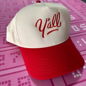May include: A cream-coloured baseball cap with a red brim and a red embroidered "Y'all" script. The cap has a red button on top. The background is pink.