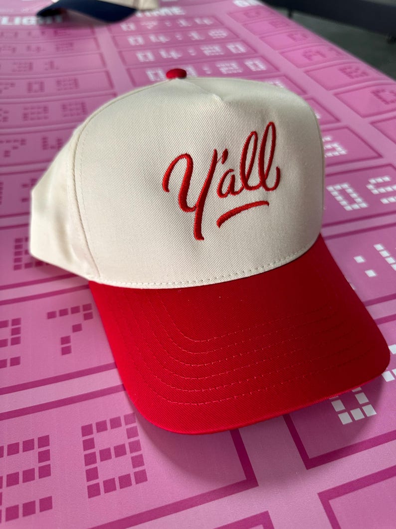 May include: A cream-coloured baseball cap with a red brim and a red embroidered "Y'all" script. The cap has a red button on top. The background is pink.
