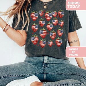 May include: Dark grey t-shirt with a pattern of strawberries designed with the American flag. The shirt is worn with blue jeans and white trainers. The text "SHIPS TODAY" and "HOUSE OF PAVAN" are visible.