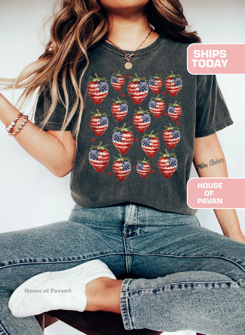 May include: Dark grey t-shirt with a pattern of strawberries designed with the American flag. The shirt is worn with blue jeans and white trainers. The text "SHIPS TODAY" and "HOUSE OF PAVAN" are visible.
