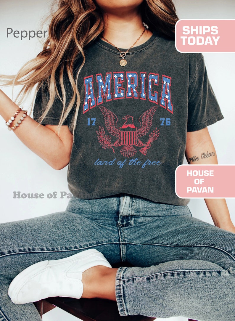 May include: A dark grey t-shirt with a vintage-style graphic of an eagle and the word "AMERICA" in blue and red lettering. The graphic also includes the numbers "17" and "76" and the text "land of the free".