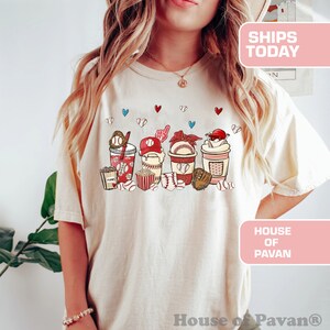 May include: A white t-shirt with a graphic of baseball-themed snacks and drinks. The graphic includes popcorn, a baseball glove, a baseball cap, a drink with a straw, and a baseball. The text "House of Pavan" is on the bottom of the shirt.