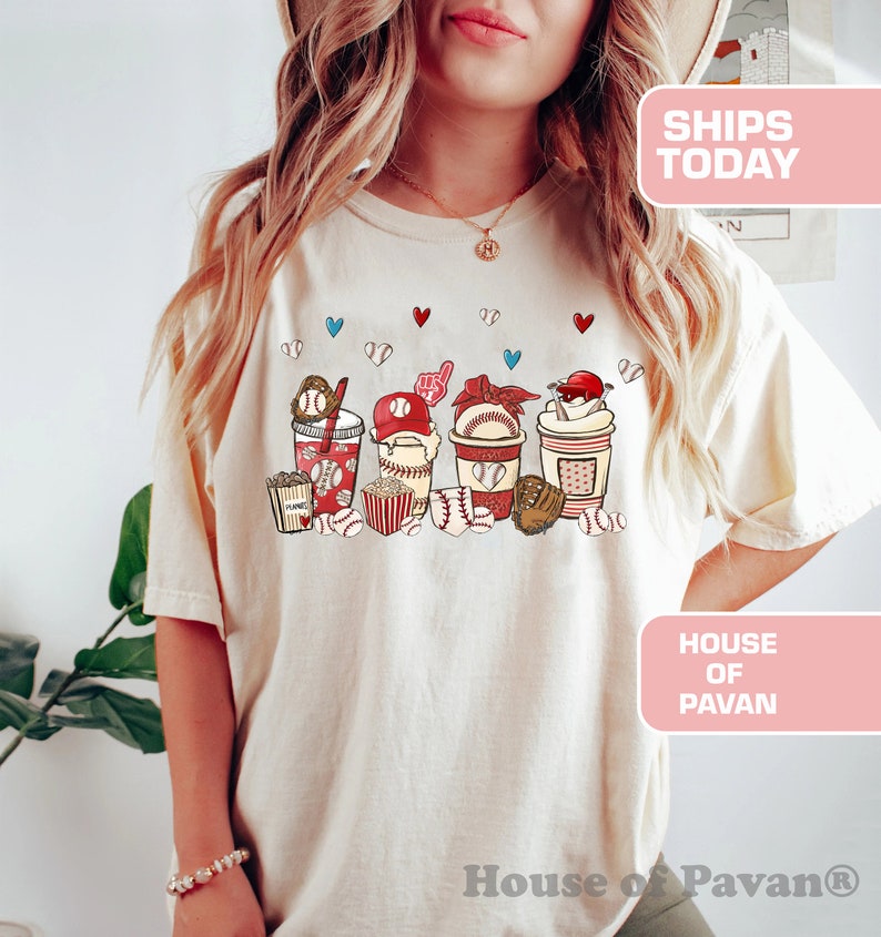 May include: A white t-shirt with a graphic of baseball-themed snacks and drinks. The graphic includes popcorn, a baseball glove, a baseball cap, a drink with a straw, and a baseball. The text "House of Pavan" is on the bottom of the shirt.