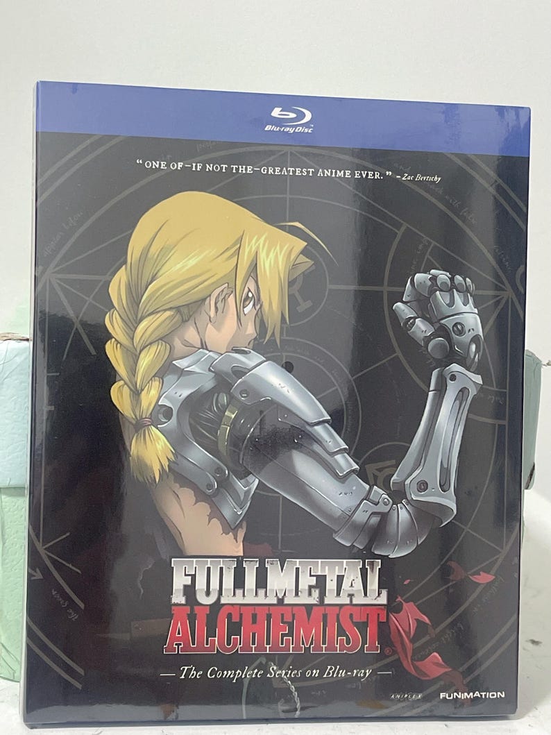 K&ouml;nnte beinhalten: Ein Blu-ray-Disc-Geh&auml;use mit der Anime-Serie Fullmetal Alchemist. Das Cover zeigt einen Charakter mit blonden Haaren und einem mechanischen Arm vor einem schwarzen Hintergrund mit einem roten und wei&szlig;en Design. Der Text auf dem Geh&auml;use lautet "Fullmetal Alchemist: Die komplette Serie auf Blu-ray".