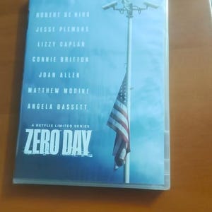 Op de afbeelding: Een dvd-hoes voor de Netflix-miniserie "Zero Day" met Robert De Niro, Jesse Plemons en anderen. De cover heeft een blauwe gradiëntachtergrond met een bewakingscamera en een Amerikaanse vlag.