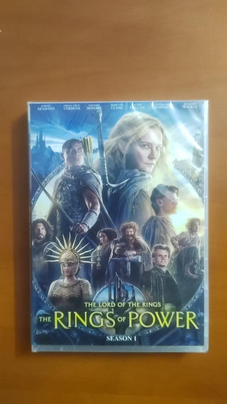 K&ouml;nnte beinhalten: DVD-H&uuml;lle f&uuml;r "Der Herr der Ringe: Die Ringe der Macht - Staffel 1". Das Cover zeigt Charaktere vor einer Fantasy-Landschaft. Der Titel ist in einer stilisierten Schriftart dargestellt.