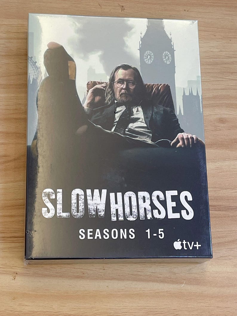 May include: A DVD box set for the television series "Slow Horses" featuring seasons 1-5. The cover art shows a man in a suit sitting in a chair with a London cityscape in the background. The Apple TV+ logo is visible.