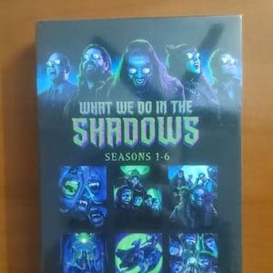 May include: DVD box set of "What We Do in the Shadows," seasons 1-6. The cover displays images of the characters and the show's title in green and blue. The box set is on a wooden surface.