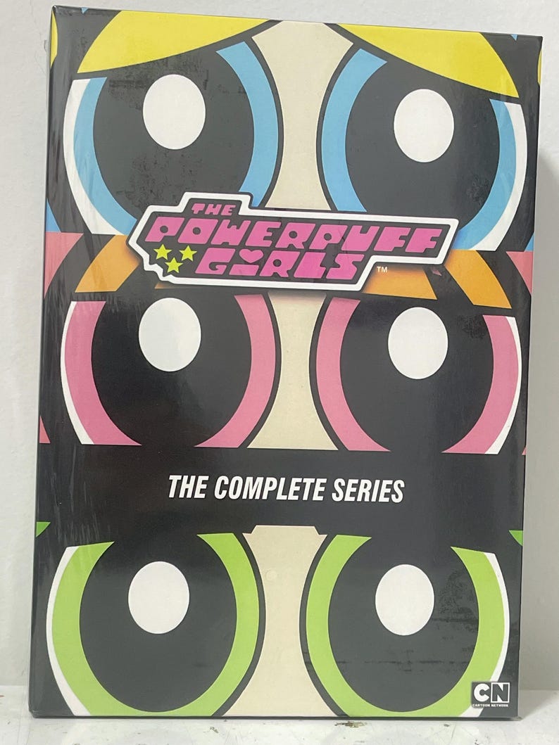 Puede incluir: Una caja negra con un dise&ntilde;o colorido que presenta tres ojos grandes. La caja tiene el texto "The Powerpuff Girls" y "The Complete Series". La caja tambi&eacute;n tiene un logotipo de Cartoon Network.