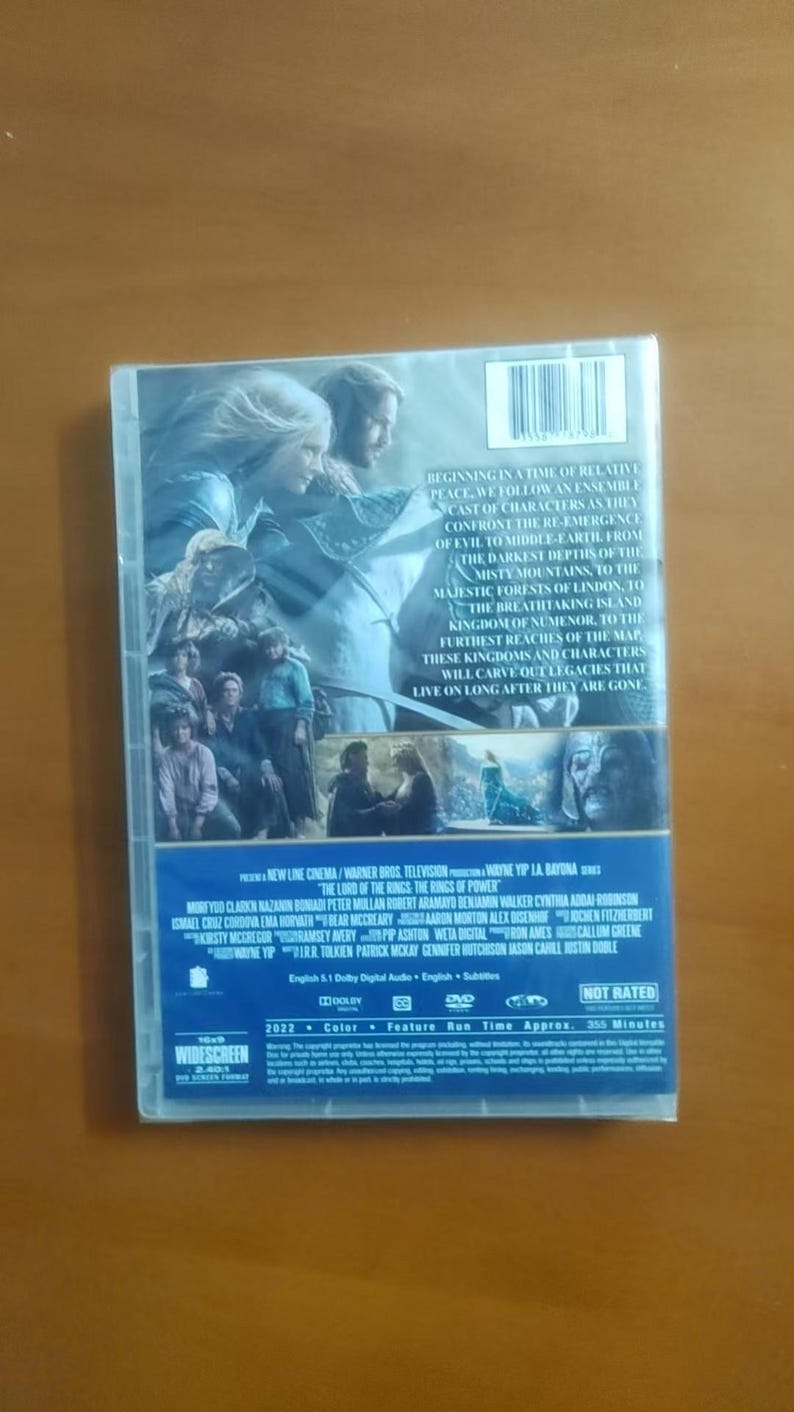 K&ouml;nnte beinhalten: Eine DVD-H&uuml;lle f&uuml;r "Der Herr der Ringe: Die Ringe der Macht" zeigt ein Landschaftsbild von zwei Figuren mit einem Berg im Hintergrund. Auf der R&uuml;ckseite der H&uuml;lle befindet sich Text, der die Handlung und die Besetzung beschreibt.