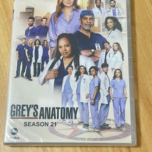 Peut inclure: Boîtier DVD pour "Grey's Anatomy" Saison 21. La couverture présente un groupe d'acteurs en tenue médicale, avec le titre de l'émission et le numéro de la saison bien en évidence. L'arrière-plan montre un paysage urbain et un coucher de soleil.