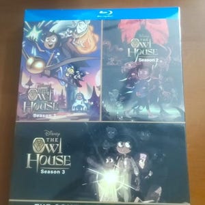 May include: A Blu-ray box set of "The Owl House" seasons 1-3. The box set features colorful artwork from the animated series, with the title and season numbers prominently displayed. The complete collection is packaged in a blue case.