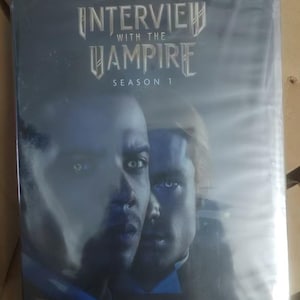 Peut inclure: DVD d'"Interview with the Vampire: Saison 1" d'Anne Rice, avec deux visages d'hommes en gros plan. La couverture sombre présente le titre en argenté. La citation "Lush and Enthralling" est en bas. Le logo AMC est visible.