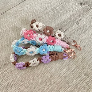 Könnte beinhalten: Fünf gehäkelte Schlüsselanhänger mit Blumendesigns in den Farben Rosa, Blau, Lila, Braun und Weiß. Die Schlüsselanhänger sind auf einer Holzoberfläche gestapelt.