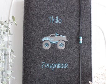 May include: A dark gray felt folder featuring an embroidered light blue monster truck graphic in the center. The name Thilo and the word Zeugnisse are embroidered in light blue above and below the truck. A gray elastic band with beads secures it.