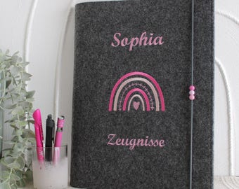 May include: A dark gray felt document folder featuring the embroidered name Sophia in pink, a pink rainbow graphic with a small heart, and the word Zeugnisse in pink. The folder is secured with a vertical gray elastic band with three beads.
