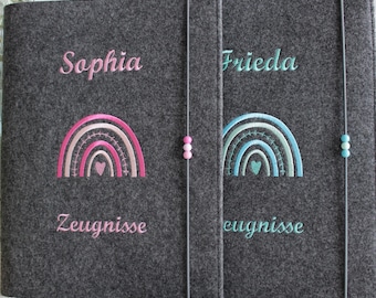 May include: Two dark gray felt folders with elastic closures featuring three small beads. The left folder is embroidered with the name Sophia, a pink rainbow, and the word Zeugnisse. The right folder features the name Frieda, a blue rainbow, and the word Zeugnisse. Two pencils lie in front.
