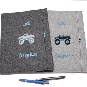 Könnte beinhalten: Zwei graue Filz-Dokumentenhalter mit gestickten Monstertruck-Designs. Einer ist dunkelgrau mit einem hellblauen Truck, der andere hellgrau mit einem schwarzen Truck. Beide haben den Namen "Emil" und das Wort "Zeugnisse" in Blau gestickt.