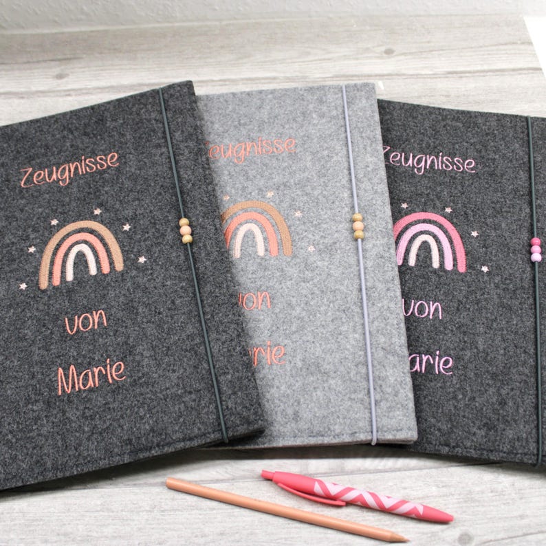 Zeugnismappe personalisiert
Drei rechteckige Filzmappen in Dunkelgrau, Hellgrau. Jede Mappe zeigt einen gestickten Regenbogen, kleine Sterne und den Text Zeugnisse von Marie in rosa Garn. Ein vertikales Gummiband mit 3 Holzperlen dienen als Verschluss.