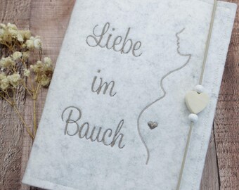 May include: A light gray felt notebook cover featuring the embroidered text "Liebe im Bauch" and a minimalist line drawing of a pregnant silhouette. A vertical elastic band with a small wooden heart bead and two white beads secures the cover.