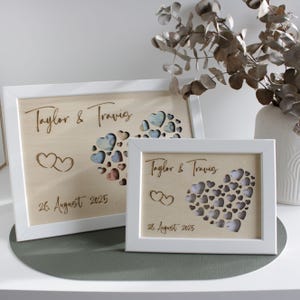 Op de afbeelding: Twee houten platen met witte randen. De platen tonen de namen "Taylor & Travis", een hartvormig ontwerp en de datum "26. August 2025". Het hartontwerp is gevuld met kleinere harten in verschillende kleuren.