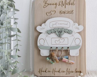 May include: A rectangular light wood plaque featuring the engraved names Bonny & Michel, the date 15.08.2026, and the German text Hand in Hand ein Leben lang. A white car cutout with the text just married, Mrs., Mr., and hearts hangs above small rolled notes.