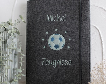 May include: A dark gray felt document folder featuring an embroidered light blue soccer ball surrounded by small stars. The name Michel is embroidered above the soccer ball, and the word Zeugnisse is embroidered below. A thin elastic band secures the folder.