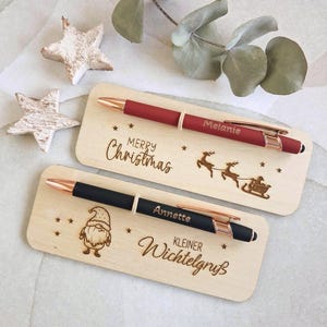 Op de afbeelding: Twee balpennen met roségouden accenten, één rood en één zwart, elk op een rechthoekige houten kaart. De rode pen is gegraveerd met "Melanie" en de kaart toont "Merry Christmas" en een rendierslee. De zwarte pen is gegraveerd met "Annette" en de kaart toont een kabouter en "Kleiner Wichtelgruß".