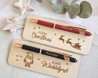 May include: Two ballpoint pens with rose gold accents, one red and one black, each mounted on a rectangular wooden card. The red pen is engraved with "Melanie" and the card features "Merry Christmas" and a reindeer sleigh. The black pen is engraved with "Annette" and the card features a gnome and "Kleiner Wichtelgruß".