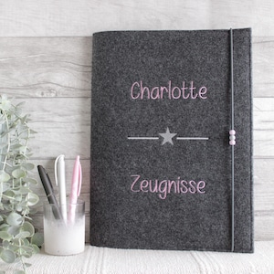 Op de afbeelding: Een donkergrijze vilten notitieboekhoes met de in roze geborduurde naam Charlotte boven een centrale zilveren ster en horizontale lijn, met het in roze geborduurde woord Zeugnisse eronder. Een elastische band met twee kleine kralen.