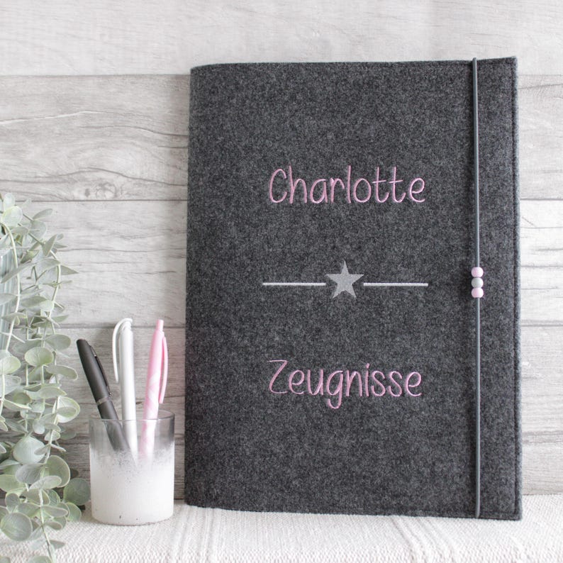 Op de afbeelding: Een donkergrijze vilten notitieboekhoes met de in roze geborduurde naam Charlotte boven een centrale zilveren ster en horizontale lijn, met het in roze geborduurde woord Zeugnisse eronder. Een elastische band met twee kleine kralen.