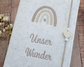 May include: A light gray felt notebook cover featuring an embroidered beige rainbow with a small heart in the center. The text "Unser Wunder" is embroidered below the rainbow. A thin elastic band with a wooden heart bead secures the cover.
