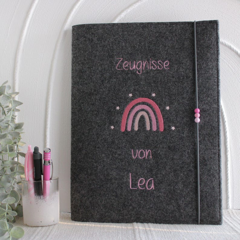 Op de afbeelding: Een donkergrijze vilten map met roze geborduurde tekst die Zeugnisse von Lea leest, gecentreerd boven en onder een roze regenboogafbeelding met kleine sterretjes. Een dun zwart elastiek met drie kleine roze kraaltjes houdt de map gesloten.